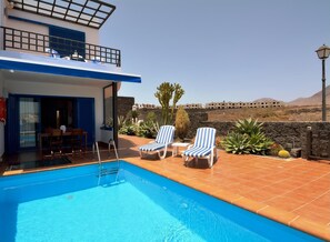 A pool area with two lounge chairs, a table, and a ladder leading to a building with a balcony. - Villa Praia Lanzarote in Playa Blanca by PVL (Playa Blanca)