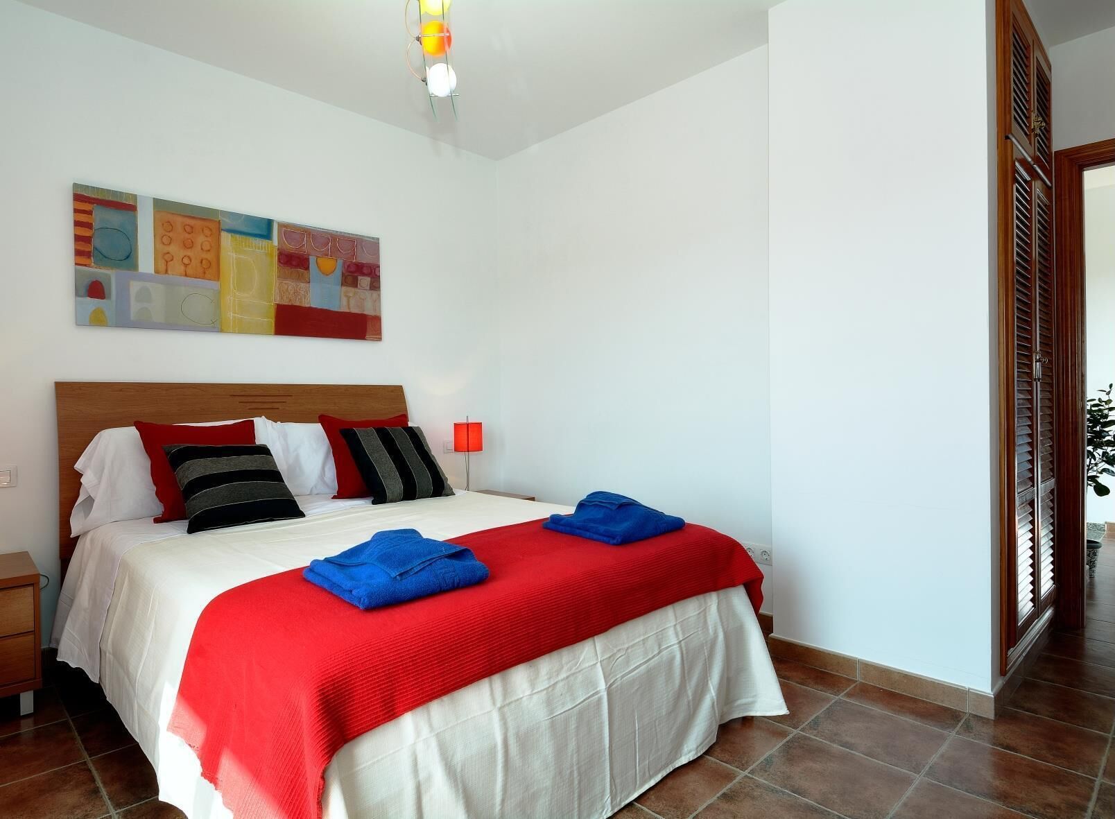 A neatly made bed with a red and white bedspread, a wooden headboard, and a red lamp beside it.