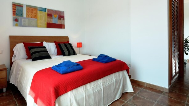 Un lit bien fait, recouvert d’une couvre-lit rouge et blanche, avec une tête de lit en bois et une lampe rouge posée sur la table de chevet.