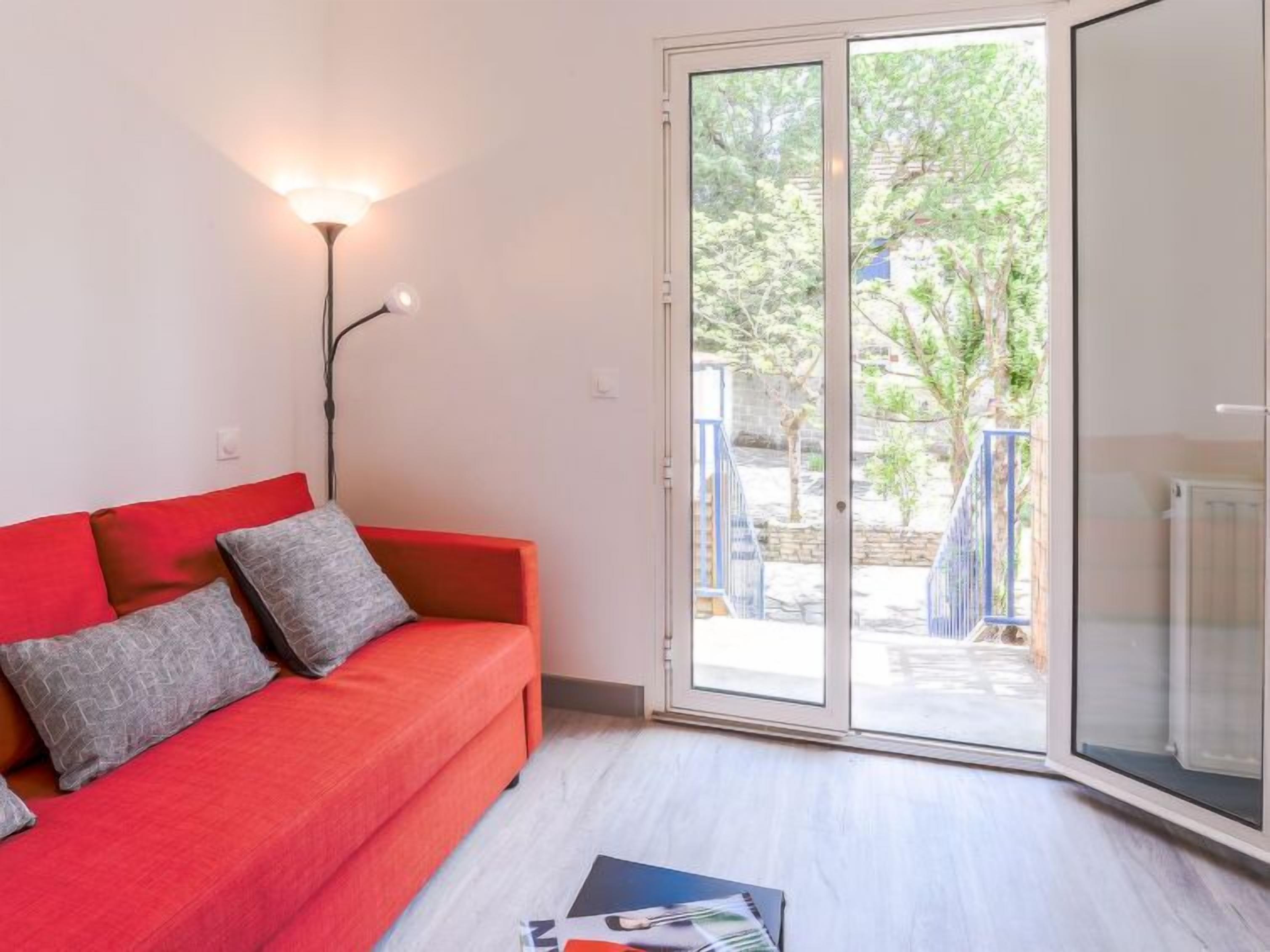 Une pièce avec un canapé rouge, un lampadaire et une porte-fenêtre coulissante donnant sur un espace extérieur doté d’une rambarde bleue.