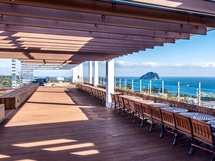 Un espacio de estar al aire libre con techo, mesas y bancos de madera, con vista al mar.