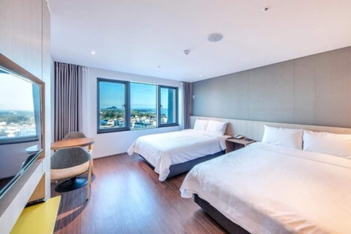 A hotel room with two beds, a large window with a city view, a round chair, and a small table.