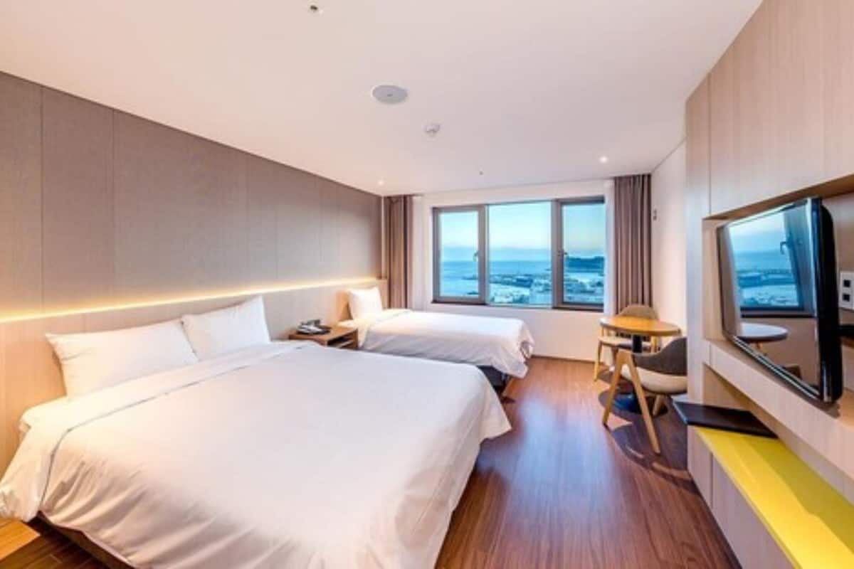 A hotel room with two beds, a TV, a small table, and a view of the sea.