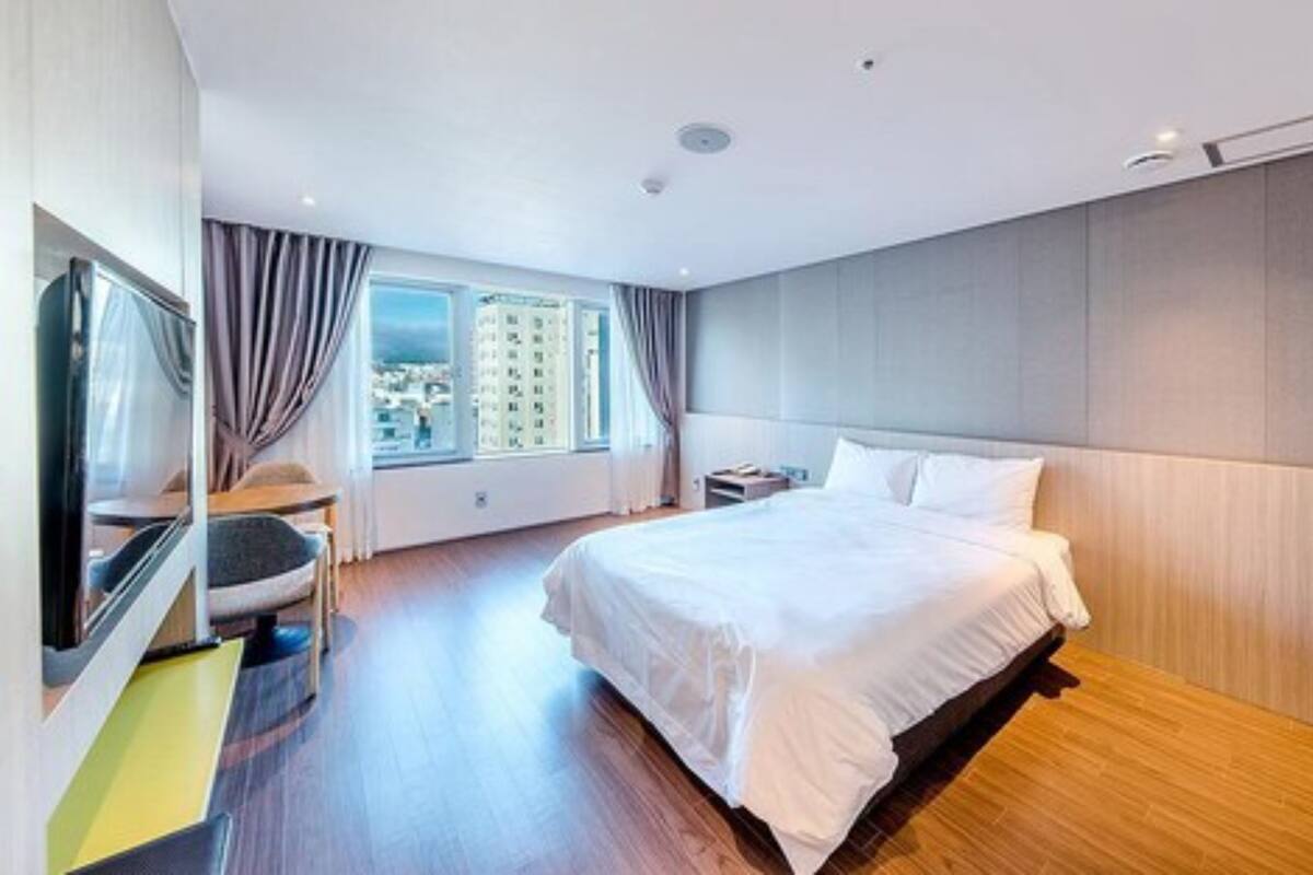A modern hotel room with a large bed, a desk, a chair, and a view of the city through the window.