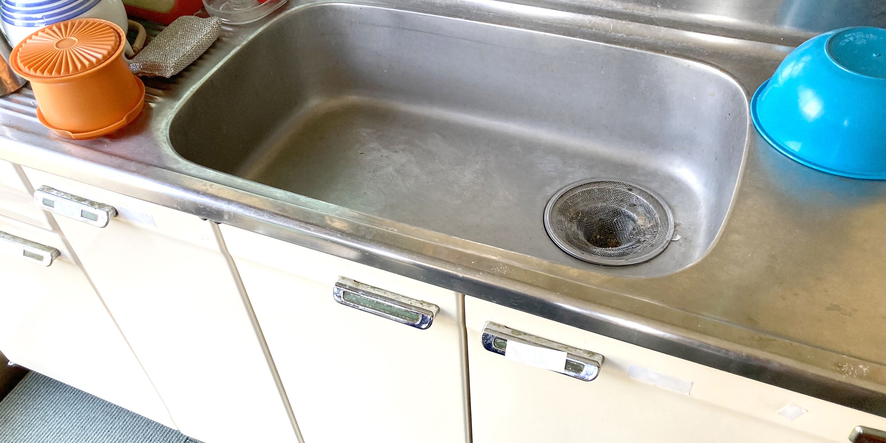 A stainless steel kitchen sink with a strainer, a blue bowl, and an orange container.