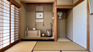 Ein traditioneller japanischer Raum mit Tatami-Bodenbelag, einer niedrigen Holzkommode, einem kleinen Regal mit Dekoration und einem gerahmten Bild an der Wand.