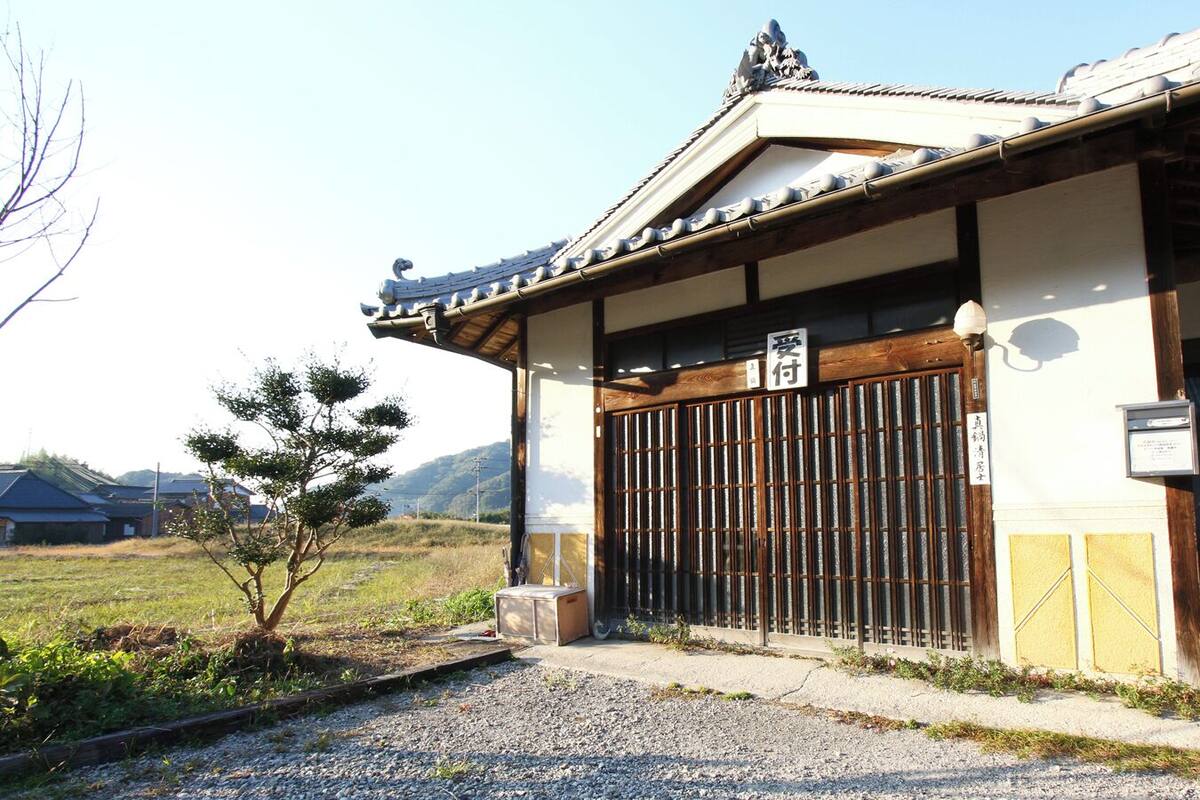 A traditional Japanese building with a wooden gate, a small tree in front, and a gravel path.