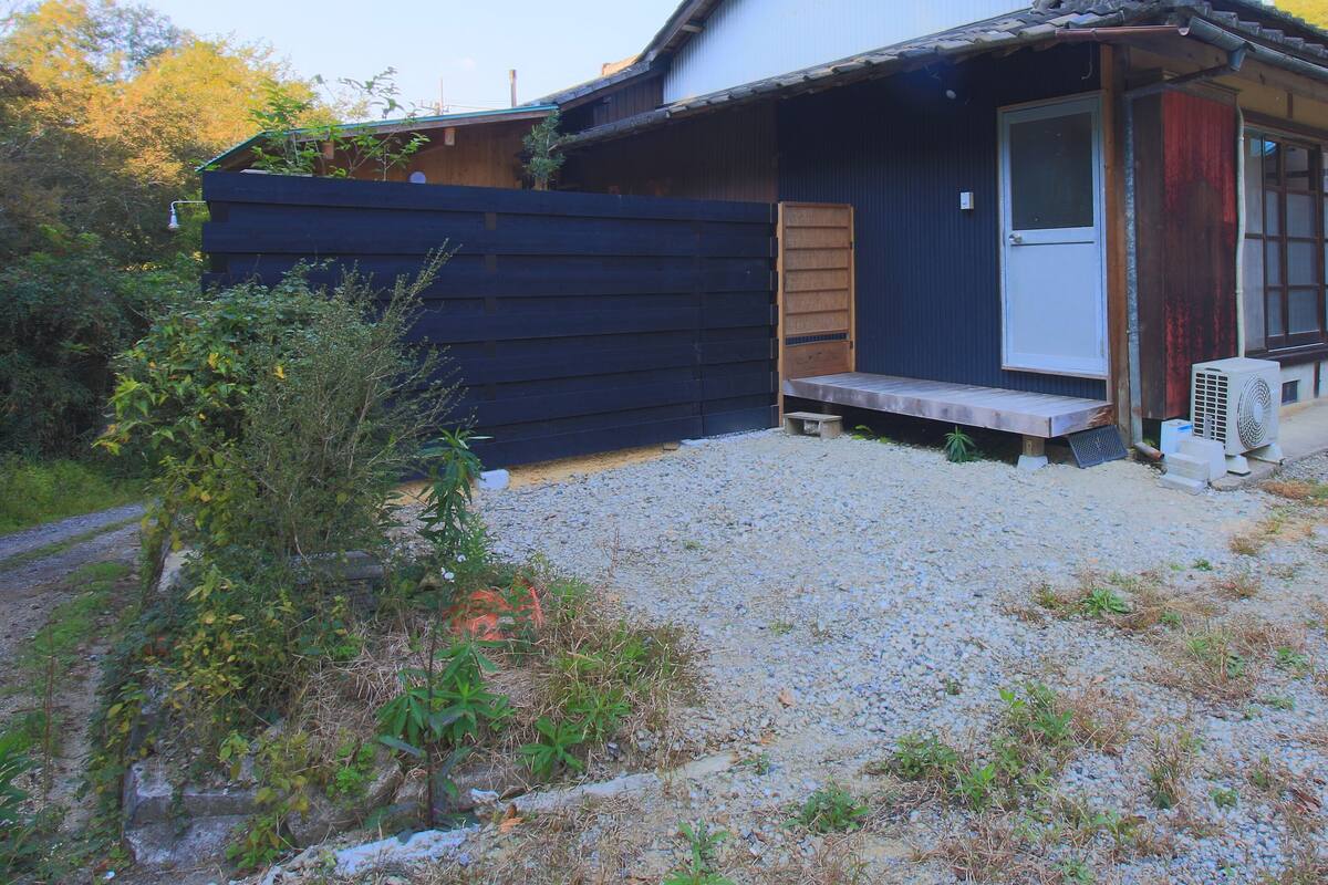 A small, rustic house with a wooden exterior, a gravel driveway, and a small porch.