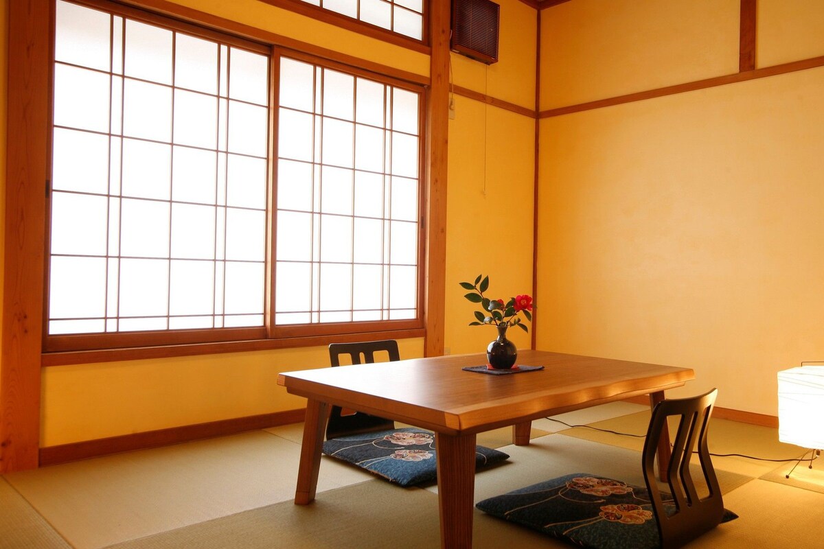 Ein traditioneller japanischer Raum mit Tatami-Böden, einem Holztisch, einer Vase mit Blumen und einem Shoji-Bildschirm.