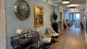 Ein Flur mit einer großen Uhr, einem Globus und einer Buddha-Statue auf Regalen. Zwei Sessel und ein Tisch mit Globus und Teekanne stehen dort. Die Wände sind mit einem gemusterten Wandpapier tapeziert, und eine Kristallleuchter ist angebracht.