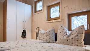Un dormitorio con cama, ropa de cama a cuadros, una bolsa colgada y dos ventanas con marcos de madera.