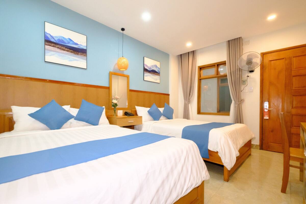 A hotel room with two beds, a wooden headboard, a large window with curtains, a wall-mounted fan, and artwork on the walls.