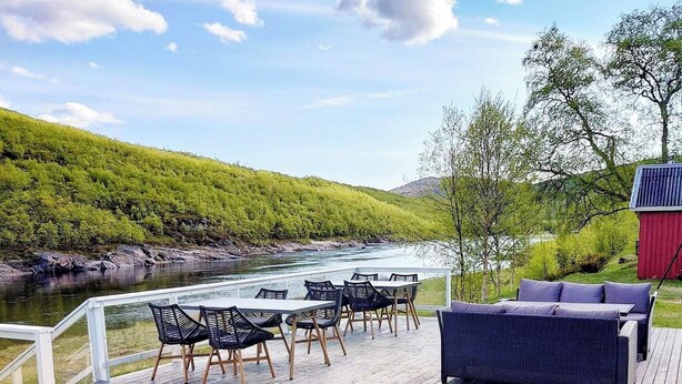En træterrasse med udendørs møbler, udsigt til en flod, og en rød bygning i baggrunden.