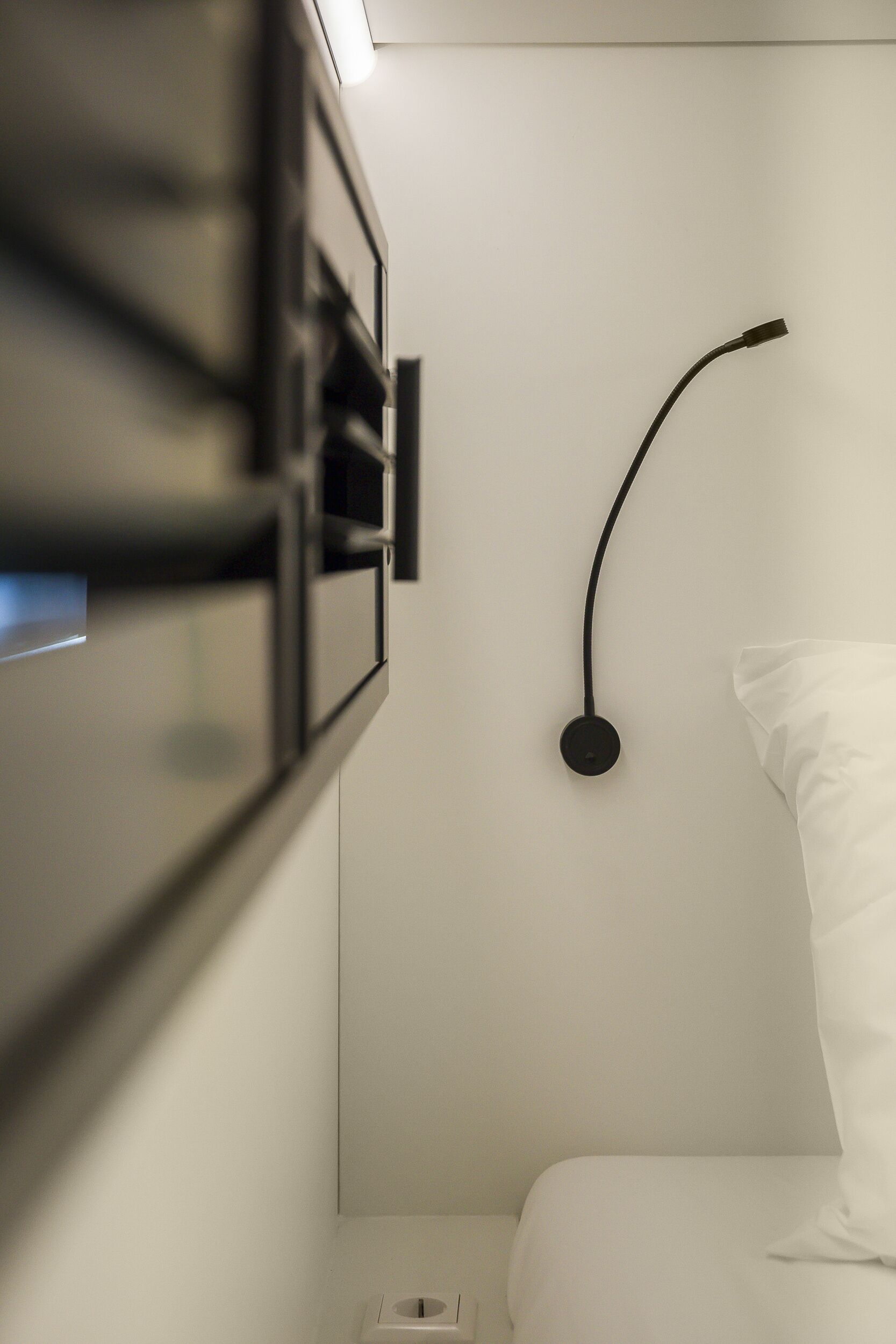 A modern hotel room with a minimalist design, featuring a bed with white bedding, a wall-mounted light fixture, and a small bedside table.