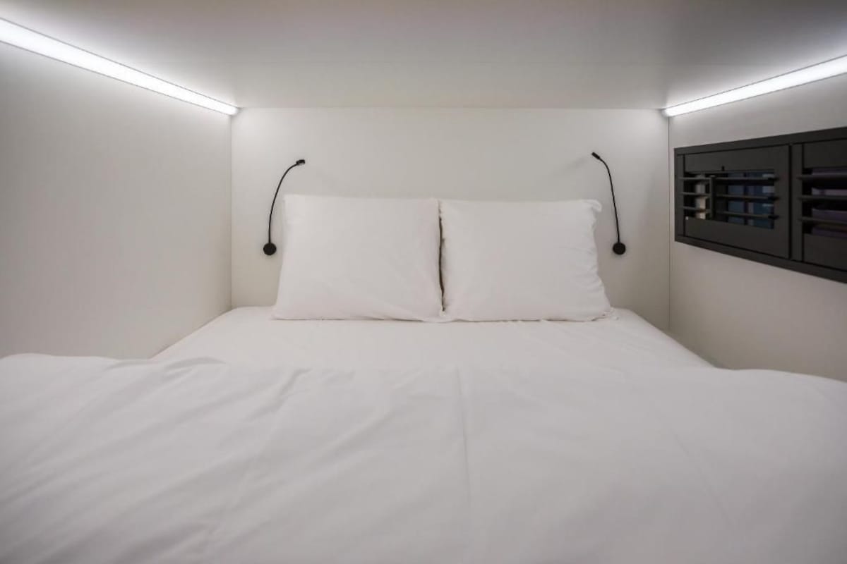 Un lit bien fait, avec du linge de lit blanc, dans une pièce aux murs blancs et dotée d’une bibliothèque intégrée sur le côté droit.