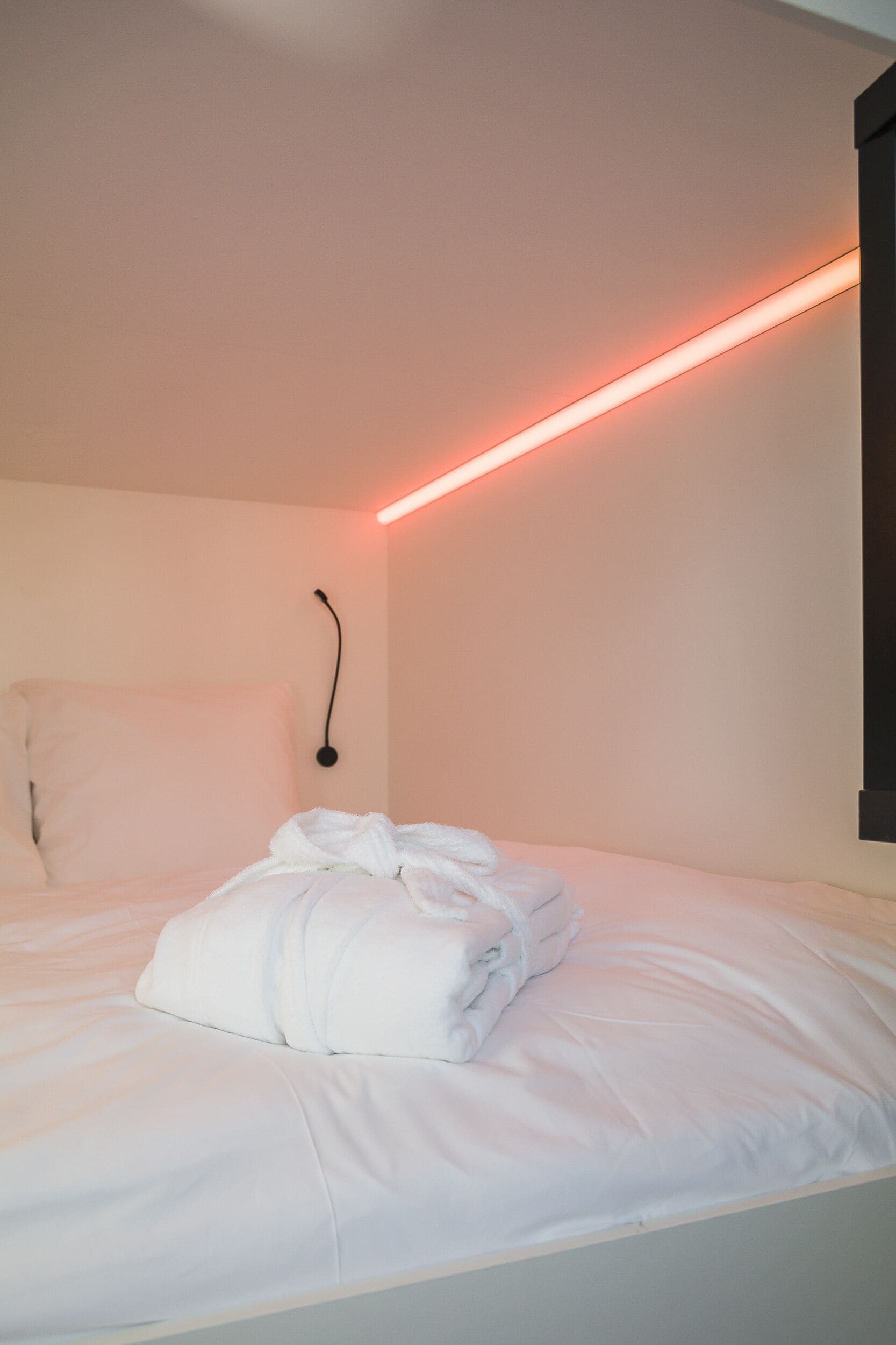 Un lit bien fait, avec du linge de lit blanc et une couverture pliée, dans une pièce dont le mur est d’un rose pâle et qui est éclairée par une bande lumineuse rouge.