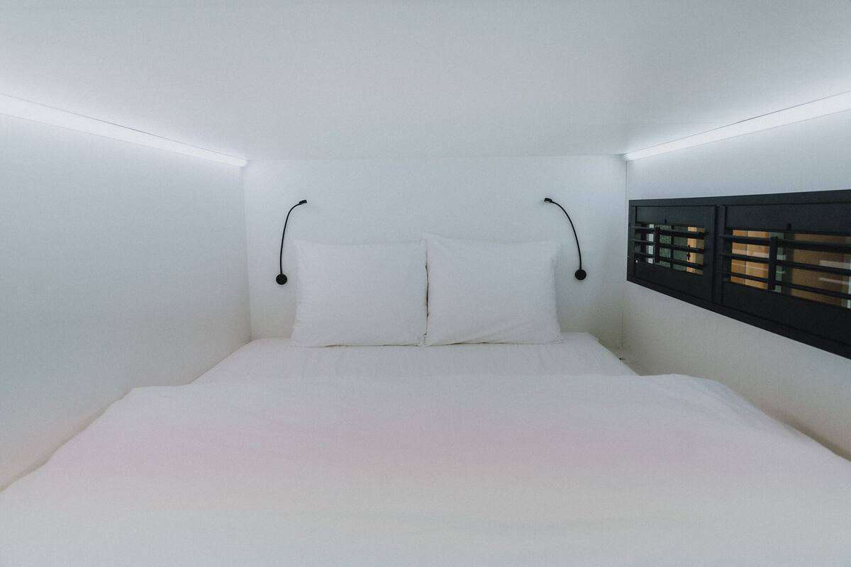 Un dormitorio minimalista con una cama, ropa de cama blanca y una ventana con persianas.
