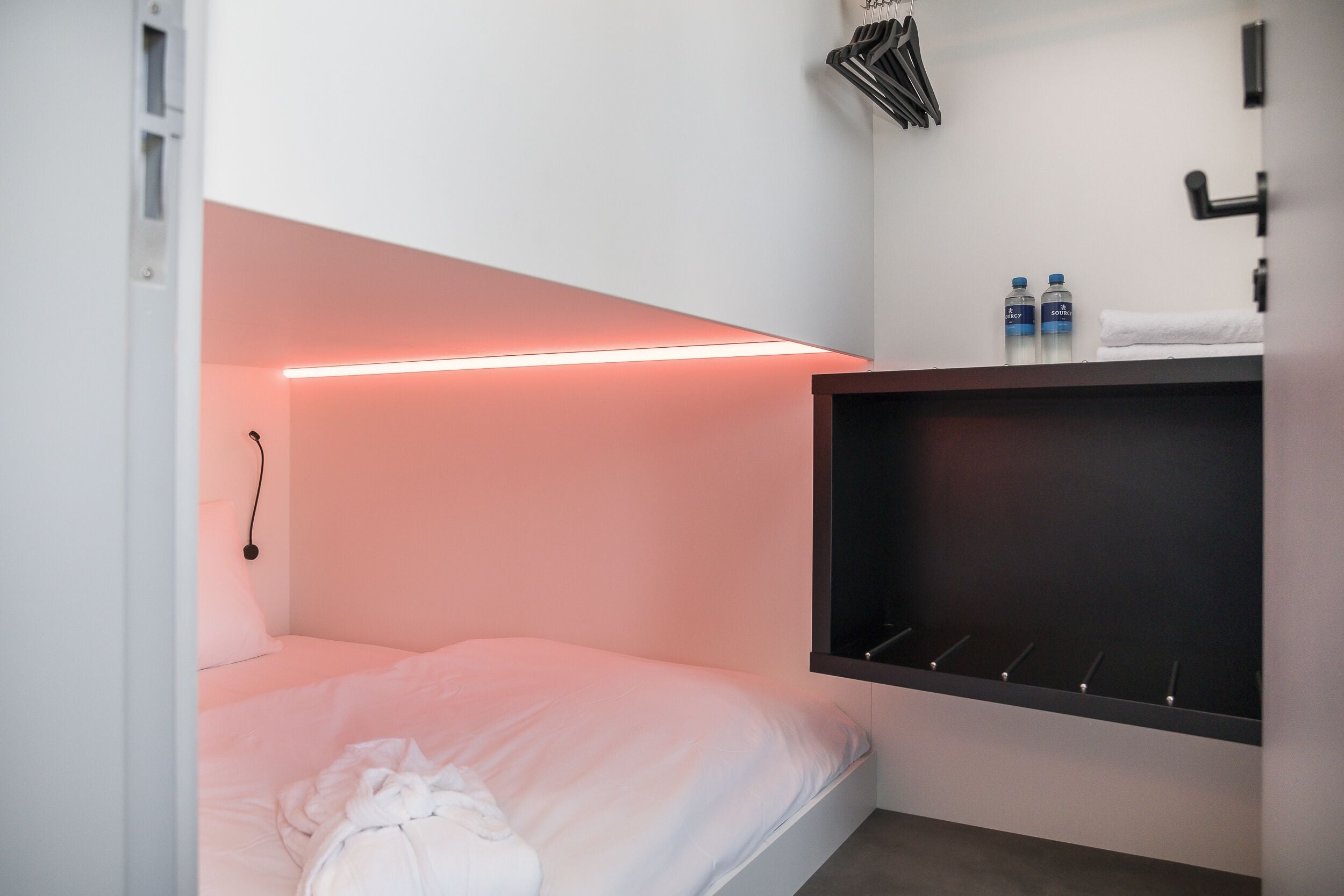 Une petite chambre à coucher moderne, avec un lit simple, une table de chevet, une étagère murale avec deux bouteilles et un porte-serviettes suspendu.