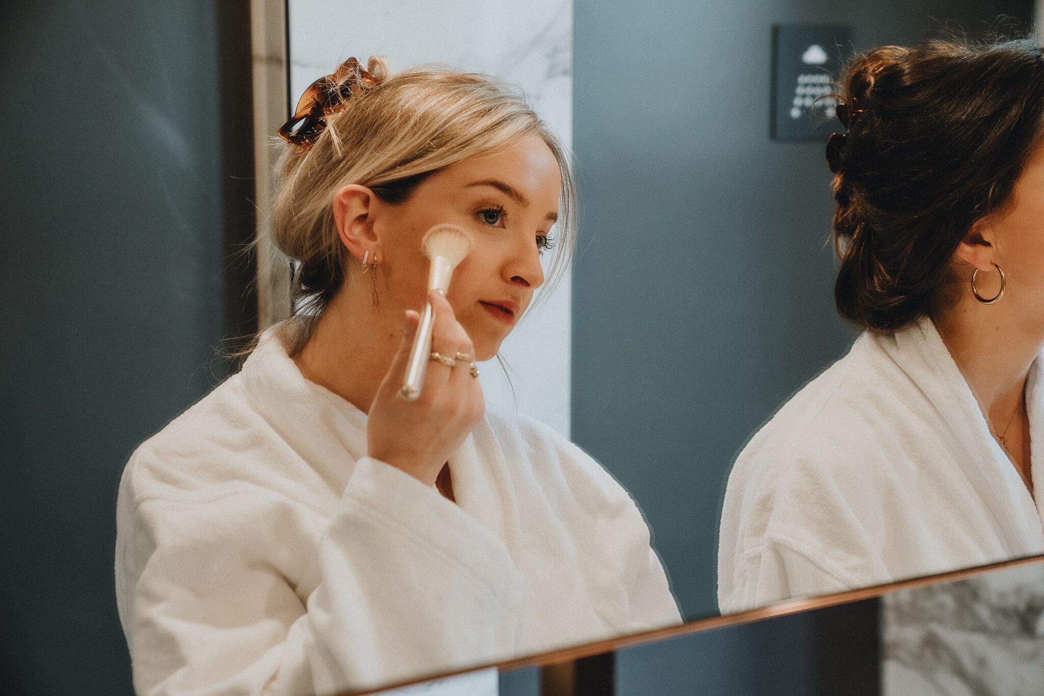 A person in a white robe applying makeup in front of a mirror.