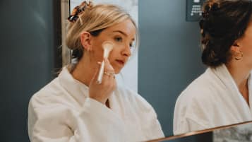 A person in a white robe applying makeup in front of a mirror.
