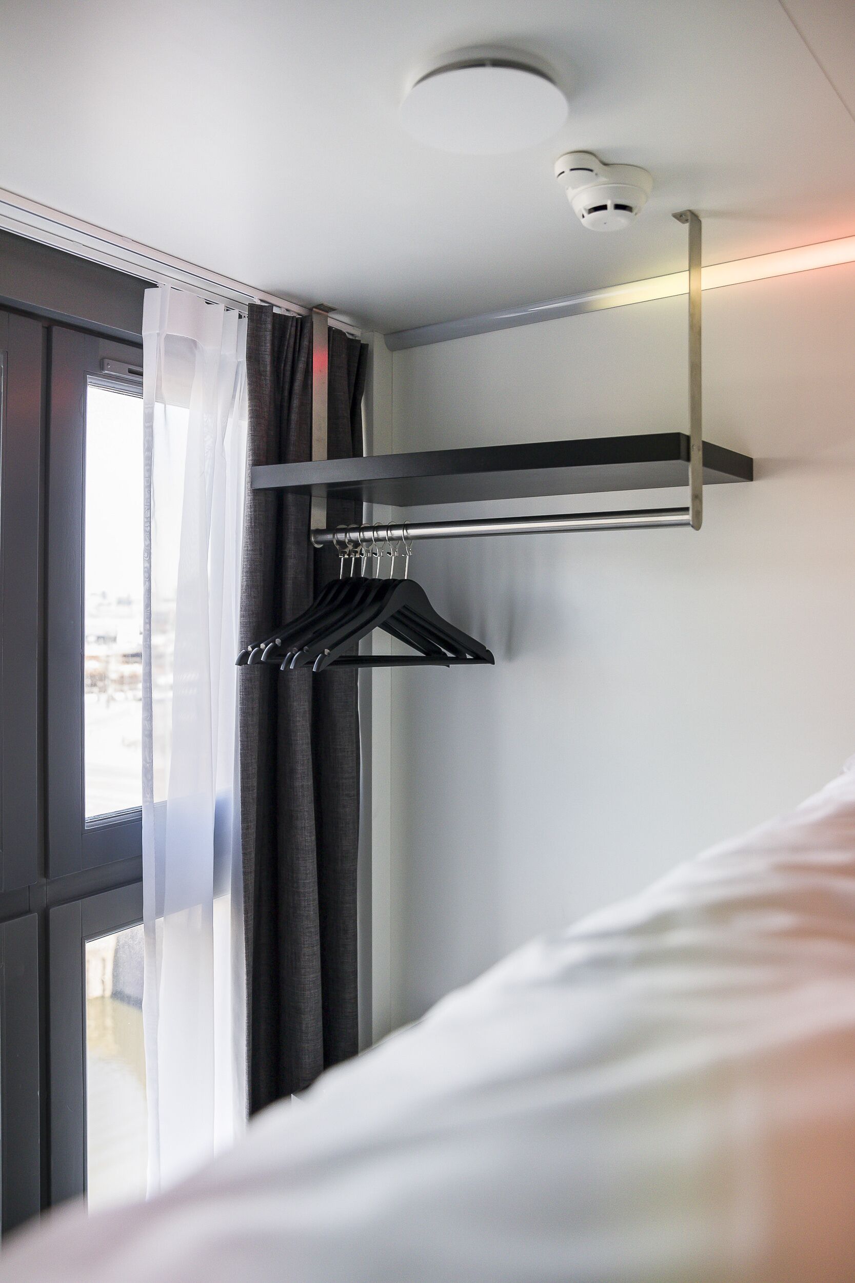 A hotel room with a bed, a window with curtains, a coat rack, and a smoke detector on the ceiling.