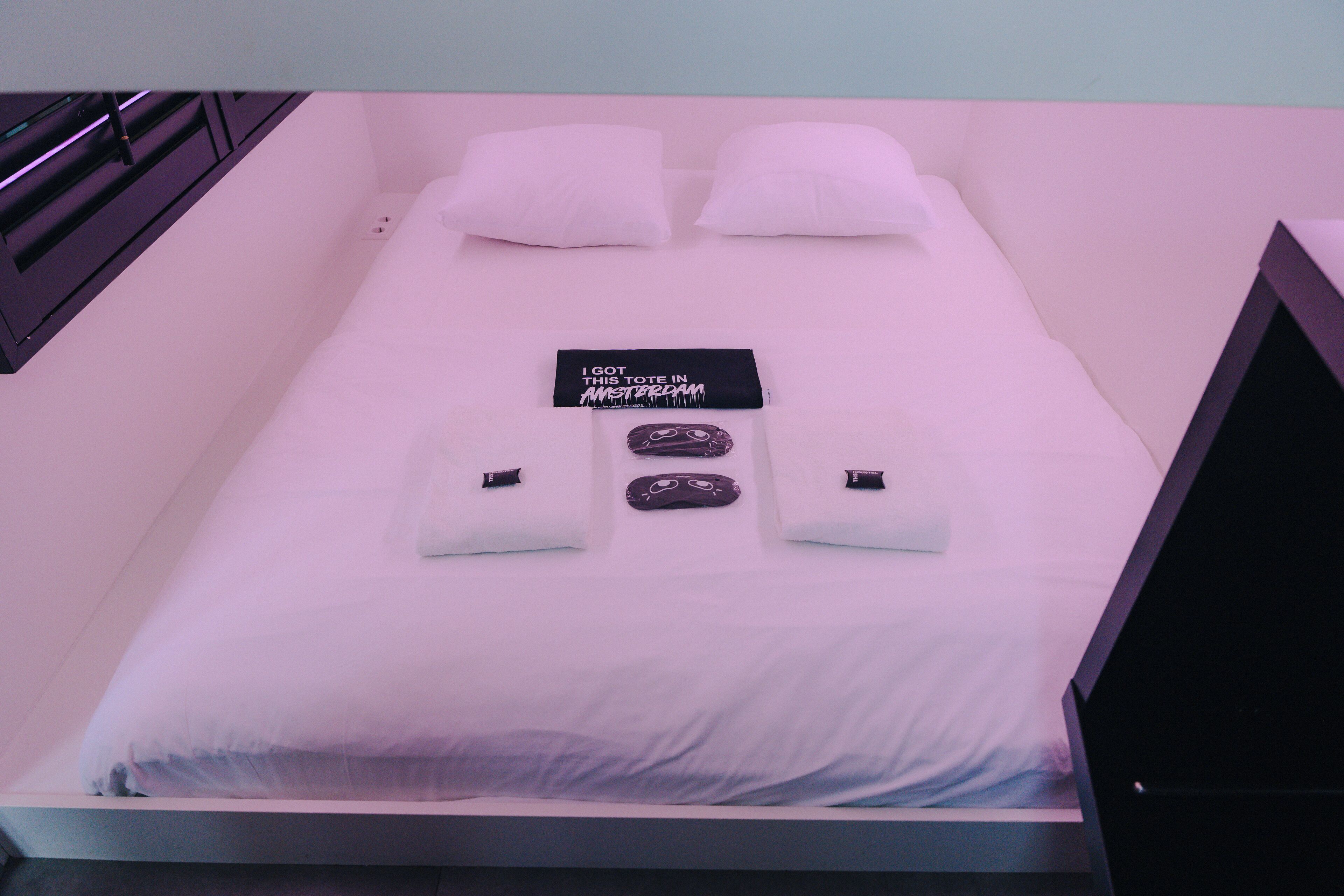 Una cama bien hecha con sábanas blancas, dos almohadas, un cartel negro sobre la cama y dos objetos pequeños negros sobre las almohadas.