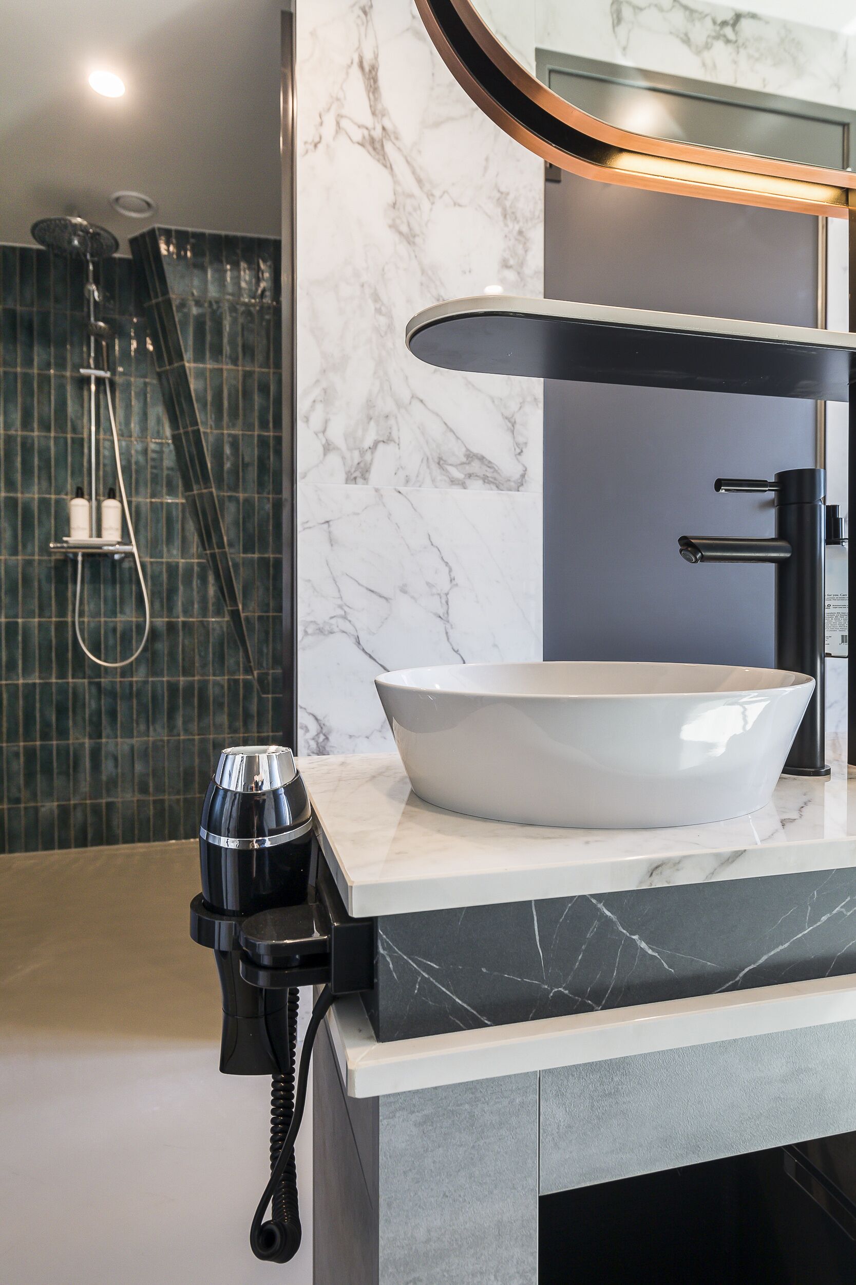 Un baño moderno con un lavamanos blanco de diseño, un cabezal de ducha manual negro y una cafetera negra sobre la encimera.