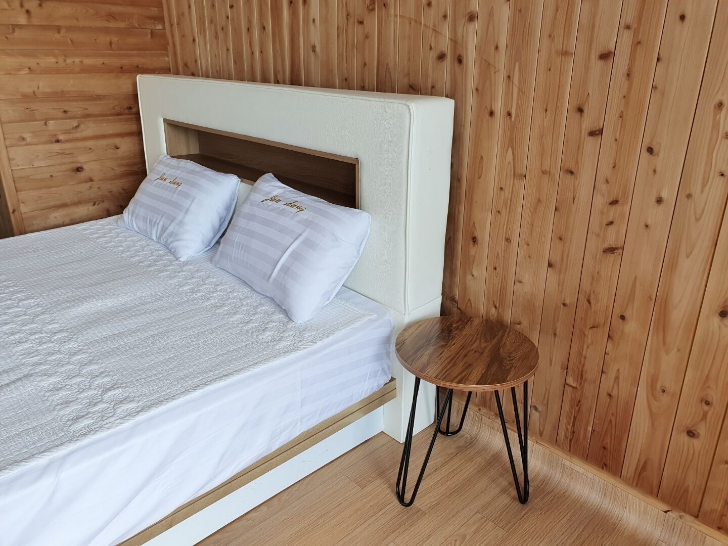 A bed with a white headboard and two pillows, a small wooden side table, and wooden panel walls.