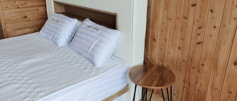A bed with a white headboard and two pillows, a small wooden side table, and wooden panel walls.
