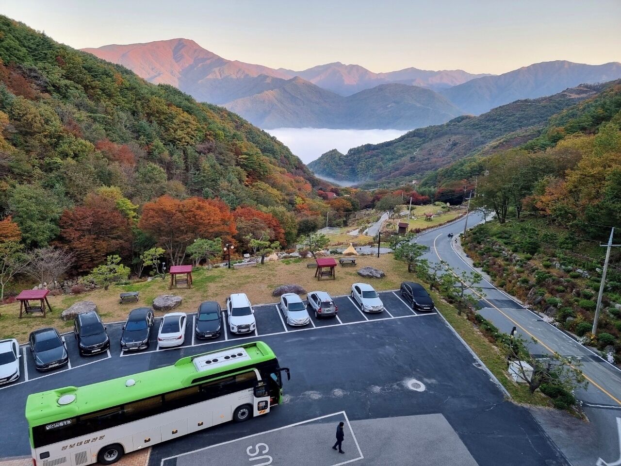 Un estacionamiento con coches y un autobús verde, rodeado de montañas y follaje otoñal.