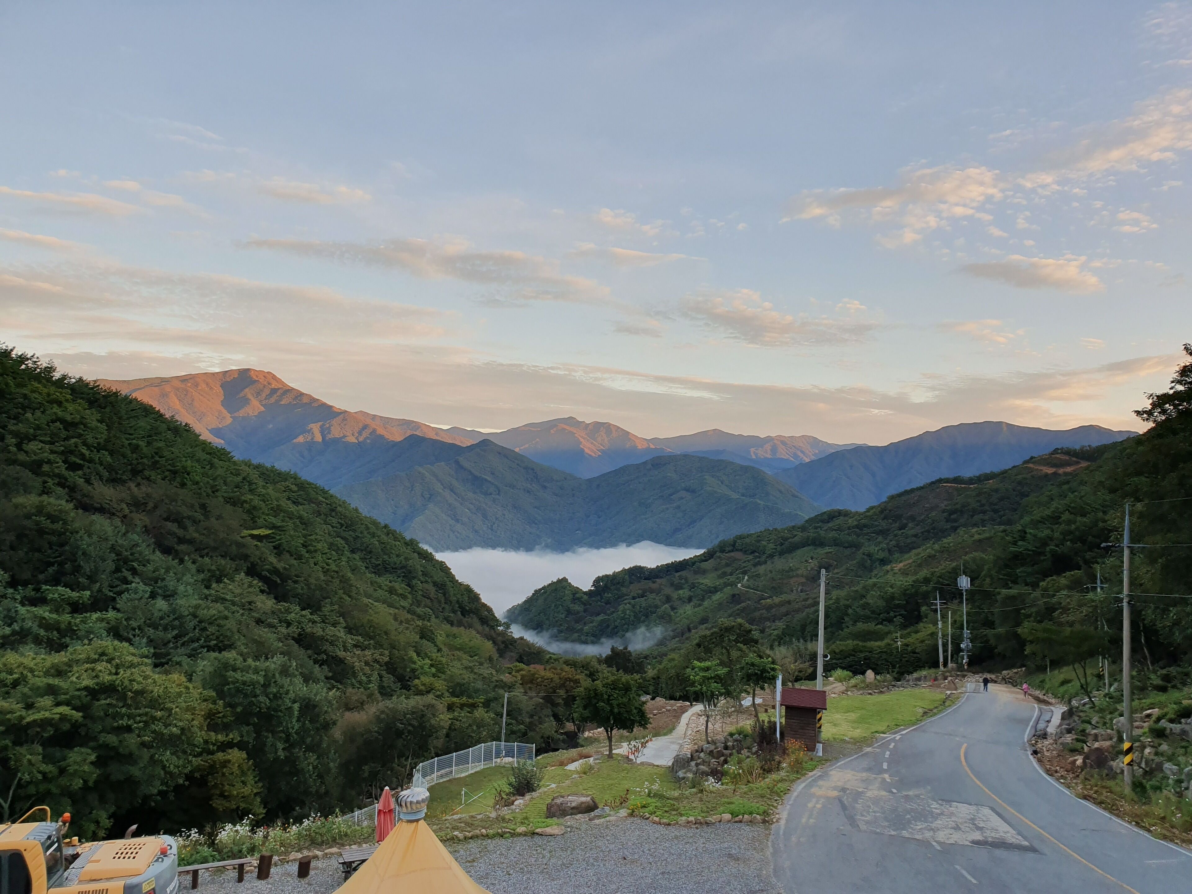 Eine kurvenreiche Straße führt durch eine gebirgige Landschaft, mit einem kleinen Gebäude und einem gelben Fahrzeug in der Nähe.