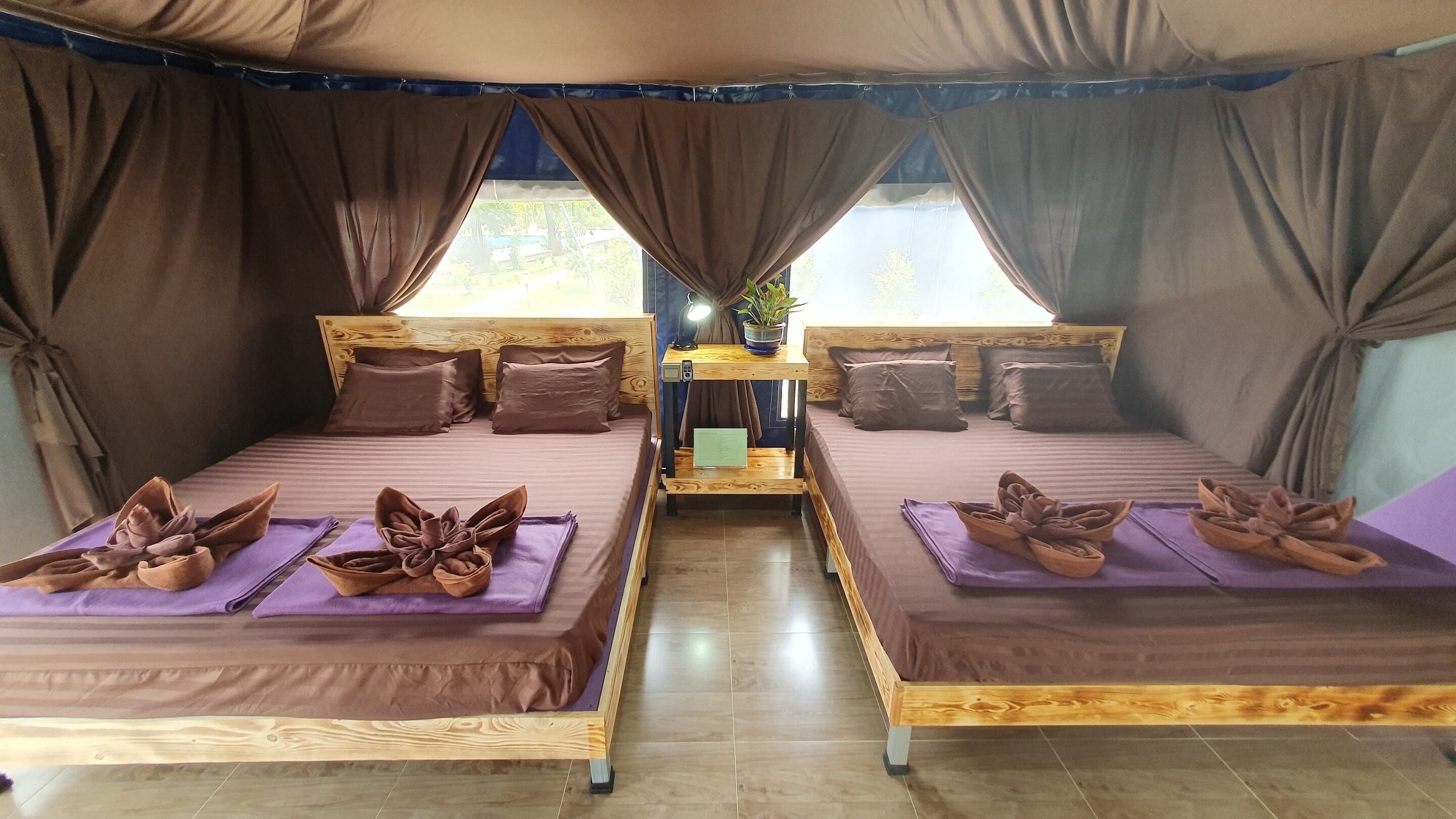 A room with two beds, each with a wooden frame and purple bedding. There is a small table with a lamp and a plant between the beds.