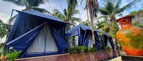 A row of blue tents with a tiled floor, surrounded by palm trees and a large decorative vase.