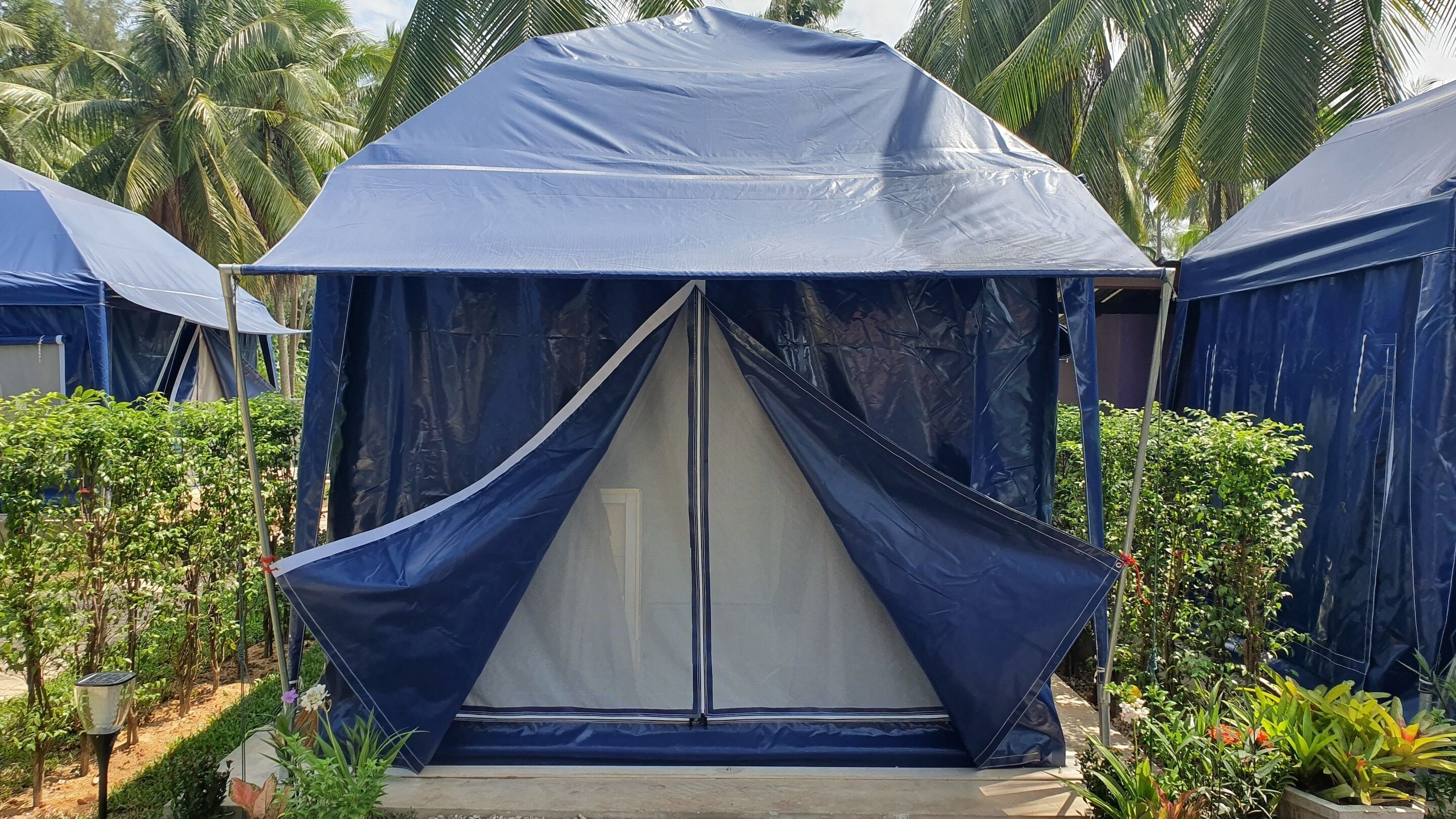 Une structure bleue en forme de tente, dotée d’une porte centrale et entourée de verdure et de palmiers.