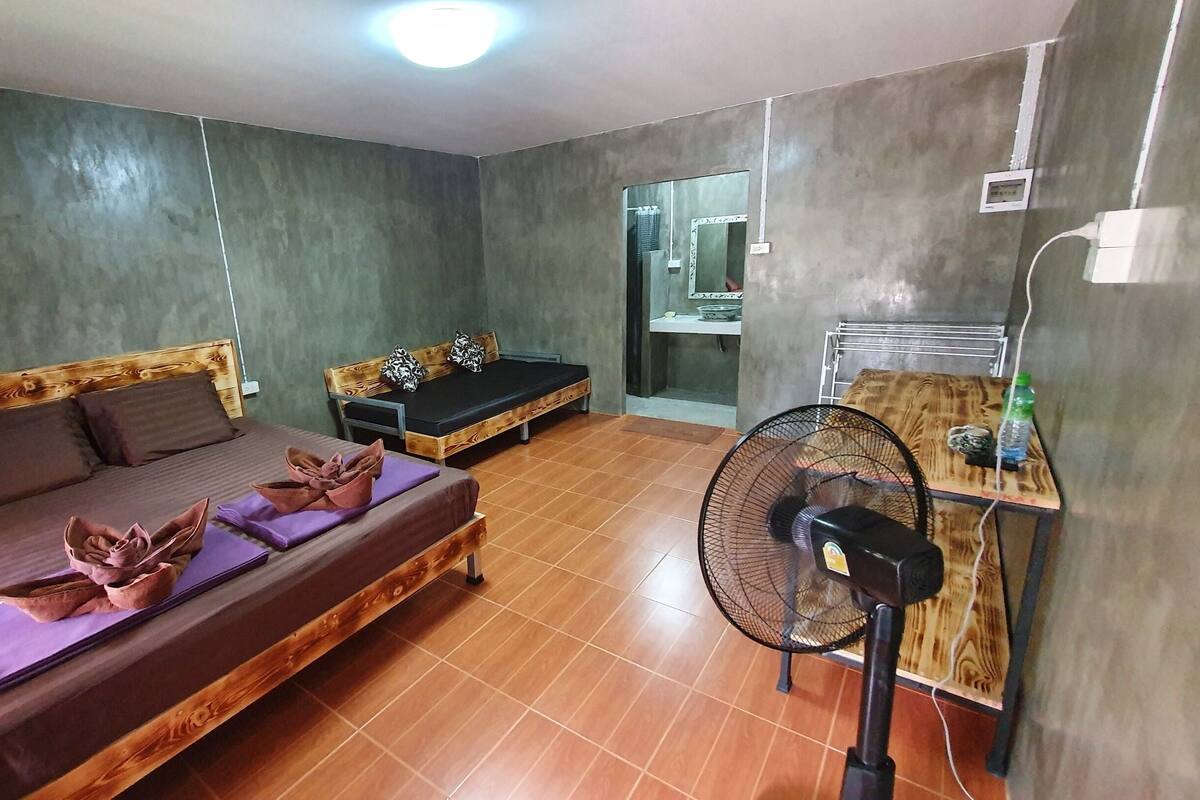A room with a bed, a smaller bed, a fan, and a shelf with a bottle.