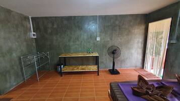 Una habitación con piso de baldosas, una mesa de madera con dos botellas, un ventilador de pie y un estante metálico.