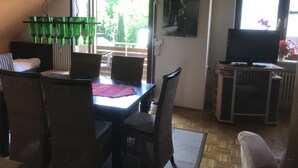 A dining area with wicker chairs, a table with a red placemat, a TV on a stand, and a window with a view of greenery. - Haus Tannenweg (Niestetal)
