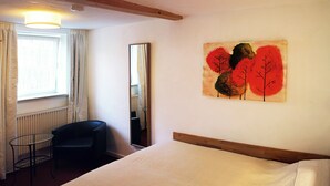 Habitación de hotel con cama, una silla, una mesa, ventana con cortinas, radiador y un cuadro en la pared.