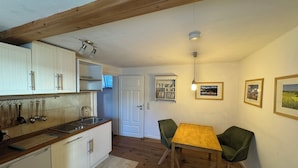 Una cocina compacta con armarios blancos, encimera de madera y un lavabo. Hay una zona de comedor con una mesa de madera y dos sillas verdes.