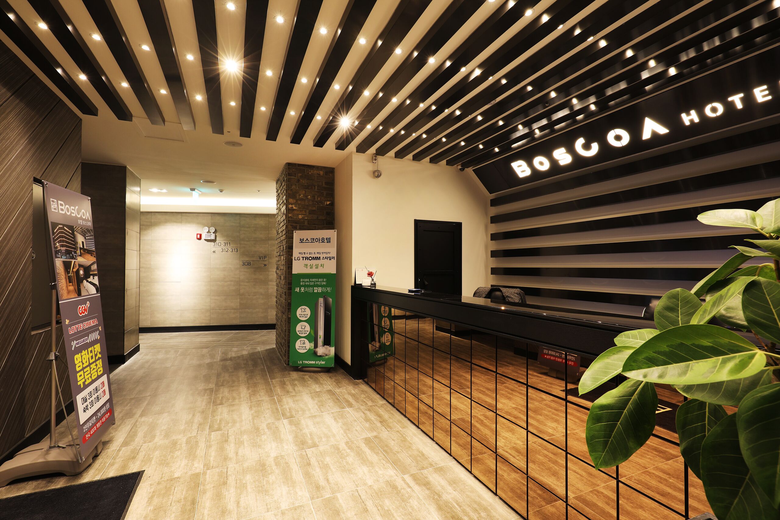 A reception area with a modern design, featuring a reception desk, a digital screen, and a sign indicating the location within the hotel.