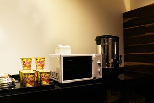 Un microondas con una cafetera y varios envases de fideos instantáneos sobre una encimera.