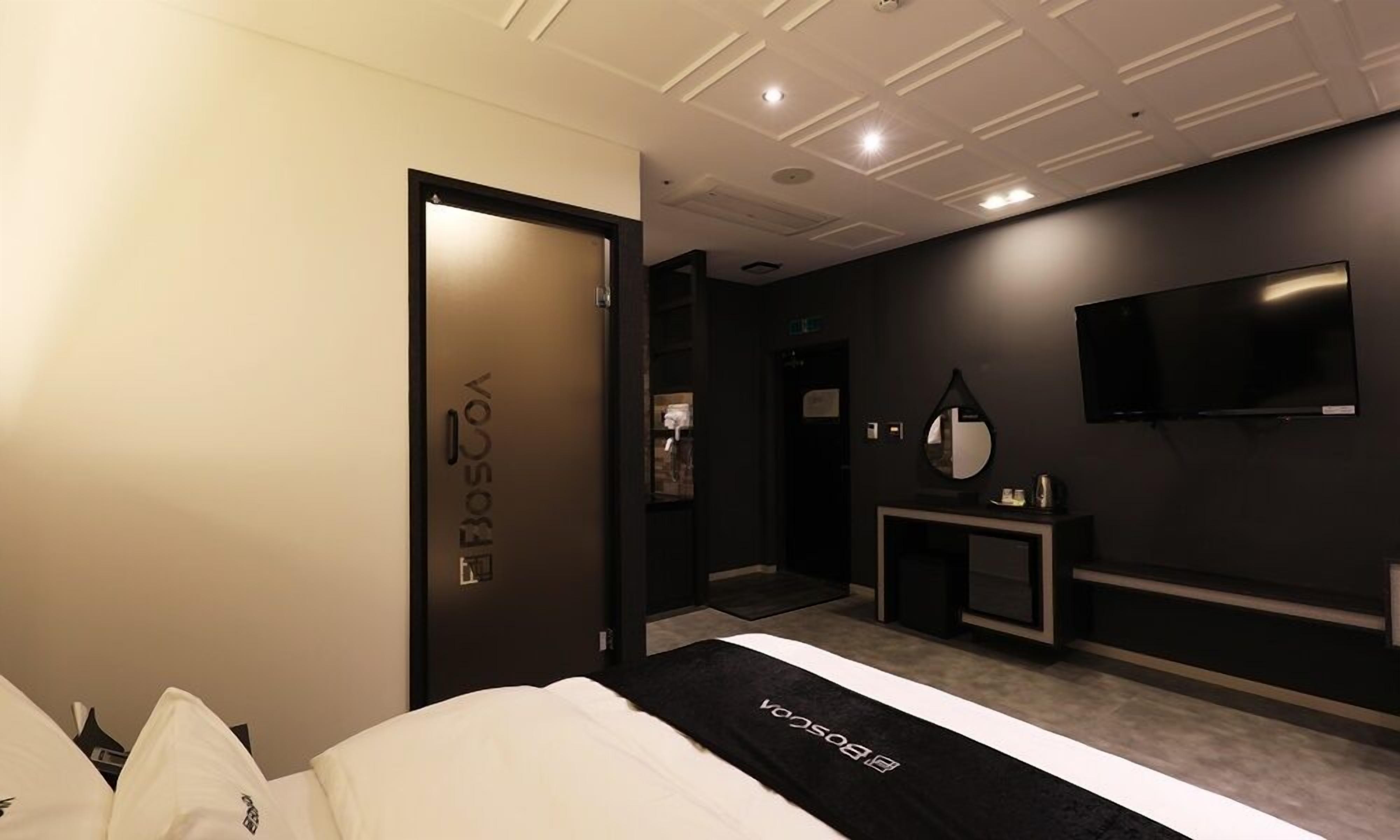 A modern hotel room with a bed, a bedside table, a mirror, a TV, and a door with a frosted glass panel.