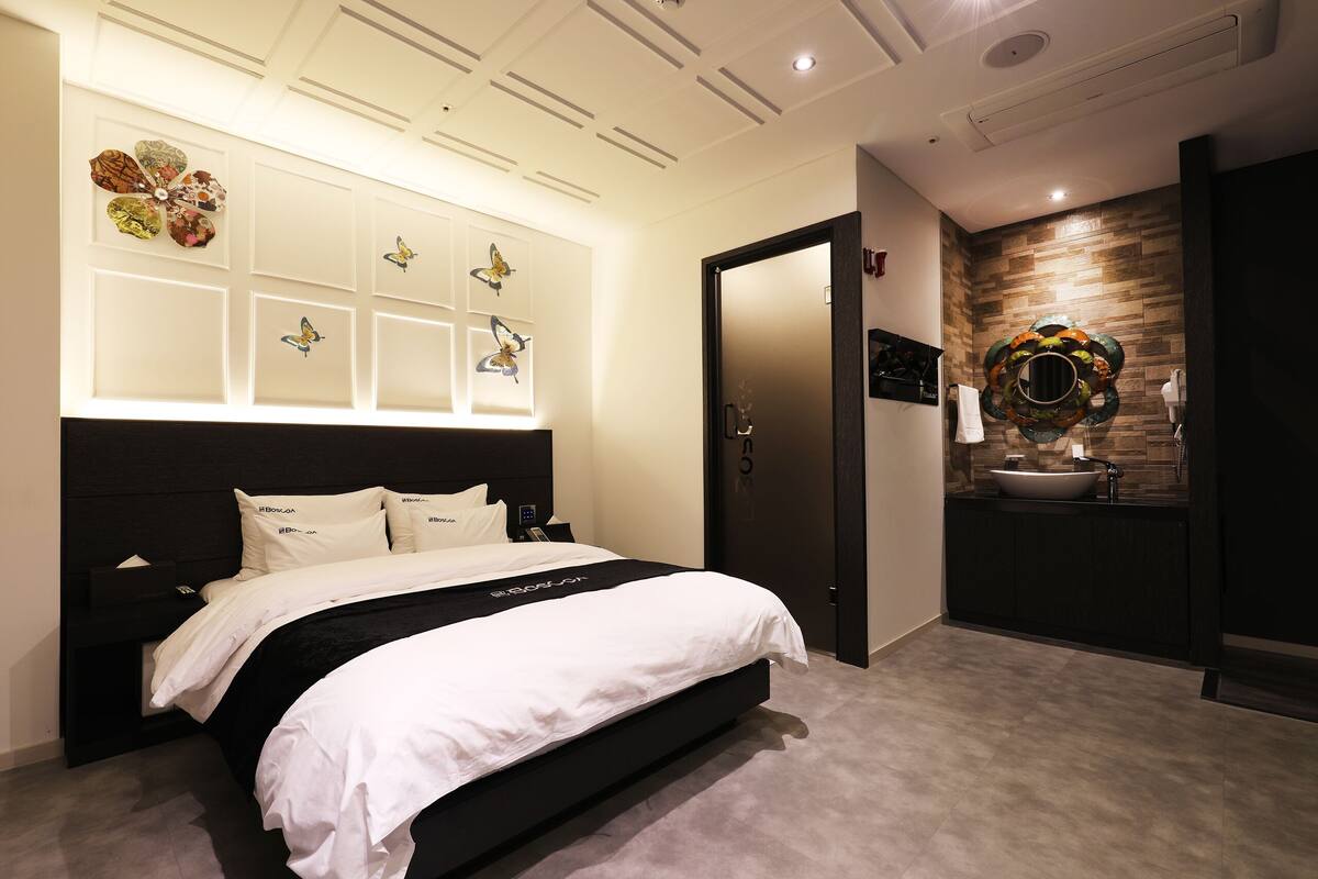 Un dormitorio moderno con una cama grande, una mesita de noche, un espejo montado en la pared y un baño con lavabo y detalles decorativos en la pared.