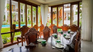 A dining area with a long table set for a meal, surrounded by wooden chairs, large windows with a view of a pool and buildings, and a staircase visible through the windows. - Caesar Phu Quoc Hotel (Phu Quoc)