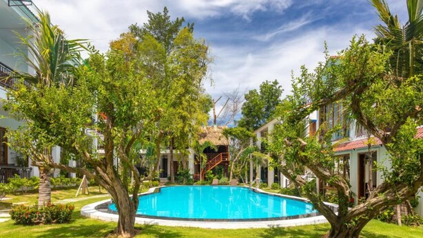 A swimming pool surrounded by lush greenery and buildings with a thatched-roof structure in the background. - Caesar Phu Quoc Hotel (Phu Quoc)