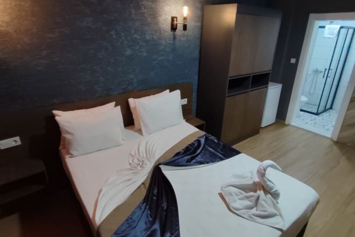 A modern hotel room with a large bed, a bedside table, a wardrobe, and a glass shower area.