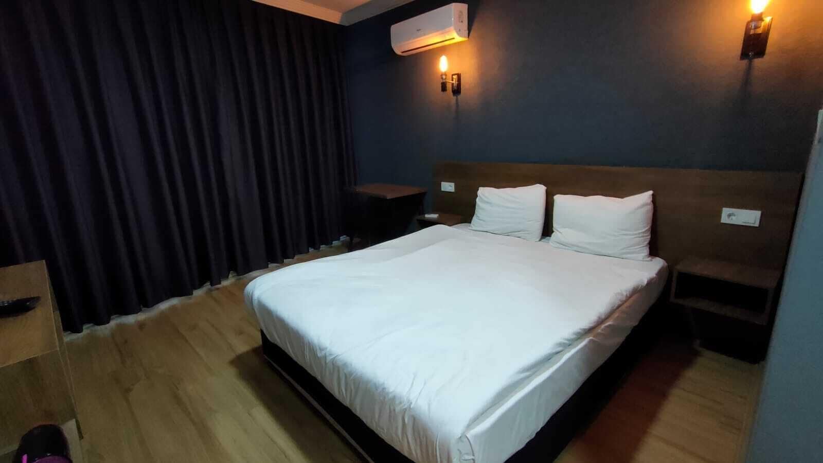 A hotel room with a large bed, a bedside table, a wall-mounted air conditioner, and a curtain.