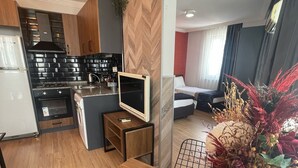 Une cuisine moderne dotée d’une cuisinière intégrée, un coin repas avec une table en bois et un salon comprenant un lit et une plante décorative.