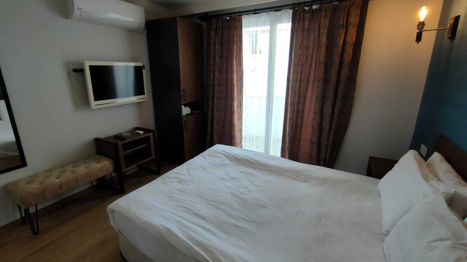 A hotel room with a bed, a television mounted on the wall, a bench, and a window with curtains.