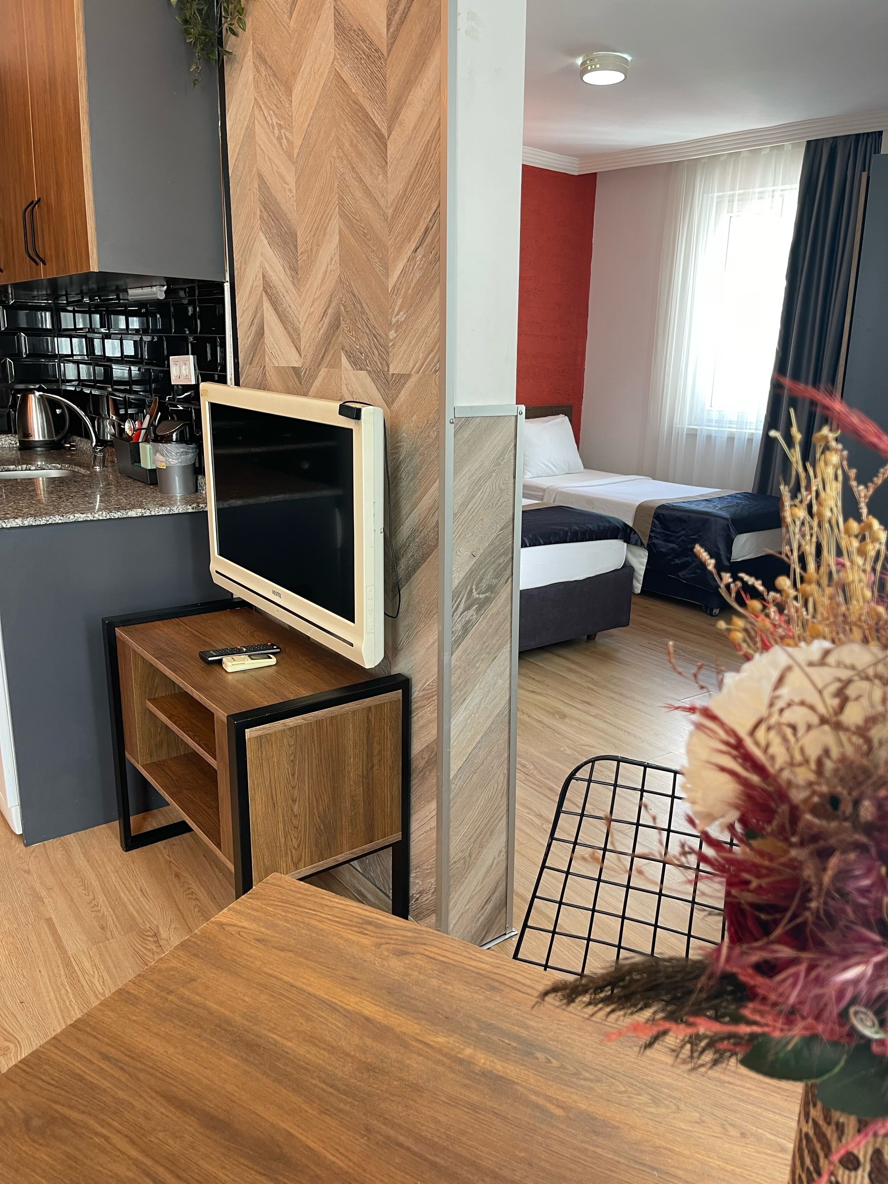 Quarto de hotel moderno com cozinha compacta, uma pequena mesa com televisão, cama e uma planta decorativa.